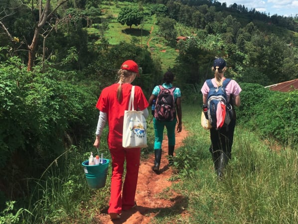 A group of volunteers carrying buckets with supplies walk on a thin dirt path through hilly Kenyan countryside.