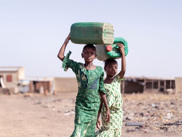 Two young girls in dresses holding large plastic water containers on their heads